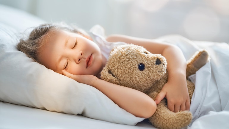 Liten jente som ligger og holder rundt teddybjørnen sin mens hun sover. En svak stråle av sollys ligger over ansiktet hennes.