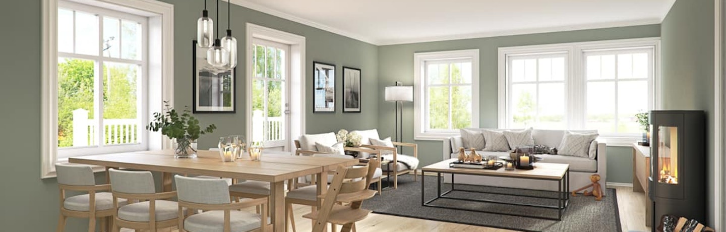 Grønnmalt stue med spisebord i lyst tre, lys grå sofa og peis i det klassiske ferdighuset Marsmorgen. Terrassedør mellom stue og spisebord og flere store vinduer som gir godt med lys.