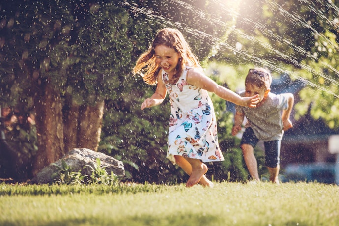 En jente og en gutt som leende løper gjennom vannstråler ute på plenen.