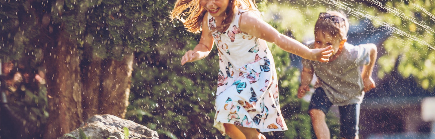 En jente og en gutt som leende løper gjennom vannstråler ute på plenen.