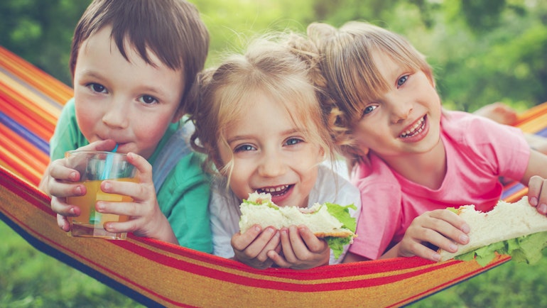 Tre smilende barn ligger i en fargerik hengekøye og smiler mot kameraet. Barna drikker saft og spiser sandwich.