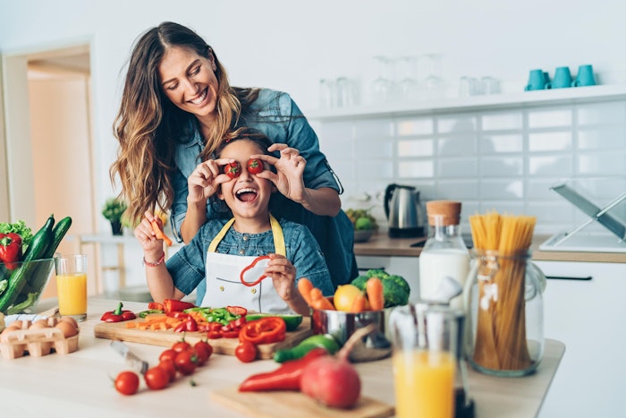 Kvinne står bak et barn og holder to tomater foran øynene på barnet. Det er grønnsaker på kjøkkenbenken og begge smiler.