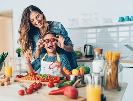 Kvinne står bak et barn og holder to tomater foran øynene på barnet. Det er grønnsaker på kjøkkenbenken og begge smiler.