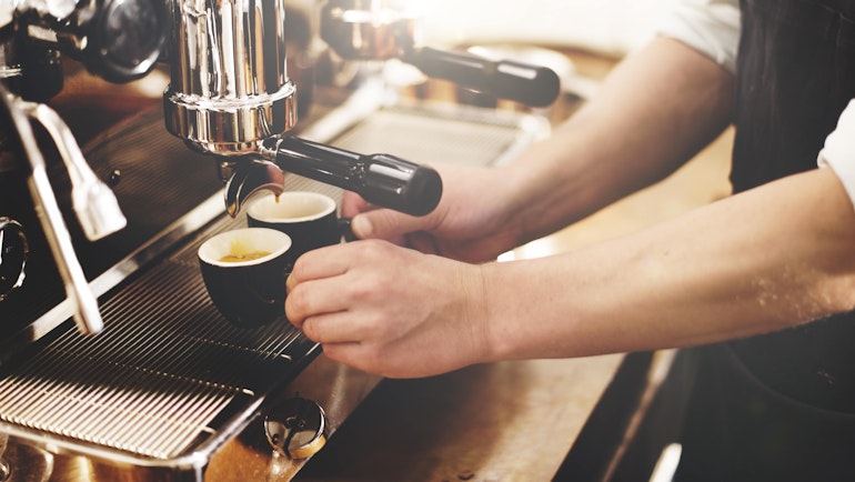 Nærbilde av kaffemaskin og en barista med forkle. Baristaen holder i håndtaket på to ferdigbryggede kaffekopper.