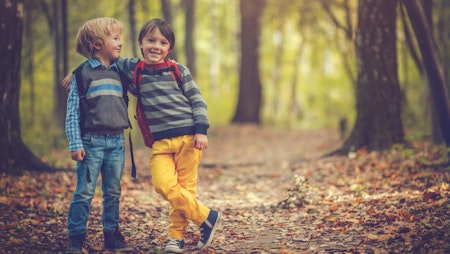 To små gutter som smiler og holder rundt hverandre. Guttene er ute i skogen og har sekk på ryggen.