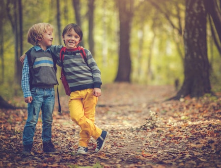 To små gutter som smiler og holder rundt hverandre. Guttene er ute i skogen og har sekk på ryggen.