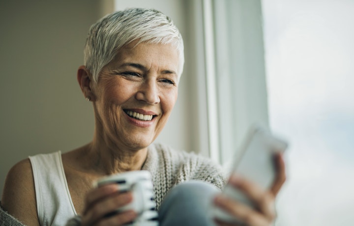Nærbilde av eldre dame som sitter ved et vindu og trykker på mobilen og drikker kaffe.