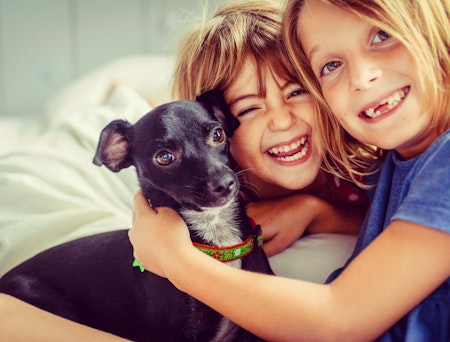 To barn og en hund sitter sammen i en seng og ser mot kameraet. De to barna smiler.