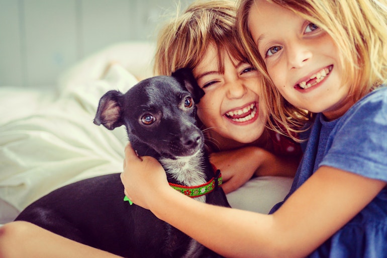 To barn og en hund sitter sammen i en seng og ser mot kameraet. De to barna smiler.