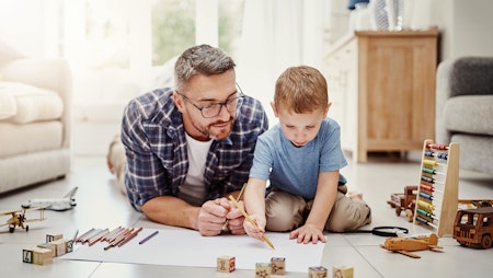 En voksen mann og en liten gutt som tegner og leker med klosser på gulvet.