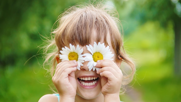 En smilende, liten jente står ute i grønne omgivelser og holder to prestekrageblomster foran øynene.